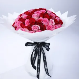 The image shows a large bouquet of red and pink roses wrapped in white paper with a black ribbon. The bouquet is placed on a white surface.