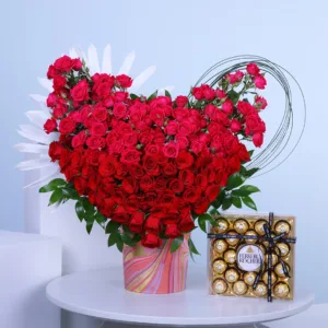 The image shows a large heart-shaped arrangement of red roses with a box of Ferrero Rocher chocolates next to it. The roses are arranged in a way that creates a full and lush heart shape, with some white flowers adding a touch of contrast. The arrangement is placed on a white table with a blue background.