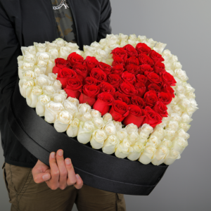 The image shows a large heart-shaped box filled with red and white roses. The red roses are arranged in the center to form a smaller heart shape within the larger one. The box is being held by a person.