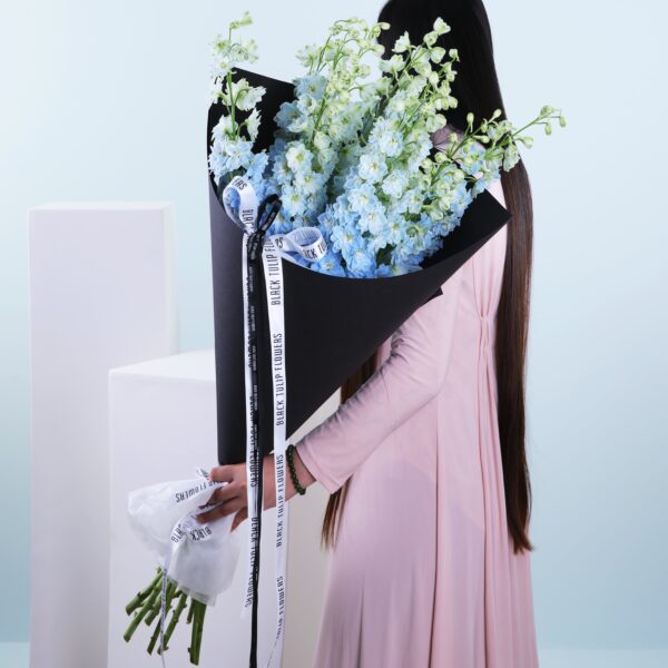 A bouquet of tall light blue delphinium flowers wrapped in black paper, held by a woman.