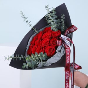 A hand-held bouquet of red roses with baby blue eucalyptus wrapped in black paper.