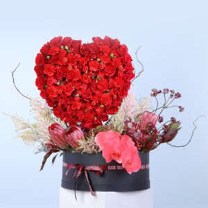 A heart-shaped bouquet made of red roses, carnations, protea, astelbi, and a phalaenopsis orchid, arranged in a black box.