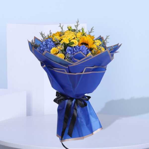 A bouquet of yellow roses, spray yellow roses, hydrangeas, delphiniums, eringium, alstroemeria, and oncidium wrapped in blue paper with a yellow ribbon.