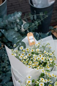 A bouquet of white daisies is wrapped in white paper and displayed on a table. The flowers have yellow centers and are arranged densely in the bouquet. There is a brown price tag attached to the bouquet with the price €5.99 written on it.