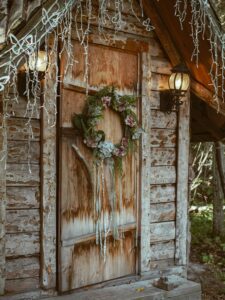 The image shows a wreath hanging on a wooden door. The wreath is made of green leaves and has a silver ribbon tied around it. There are also small silver ornaments on the wreath. The door is made of dark wood and has a diagonal pattern.