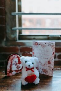 The image shows a small teddy bear holding a red heart. It is sitting on a wooden table in front of a window with a brick wall behind it. There is also a Valentine's Day card with a heart on it.