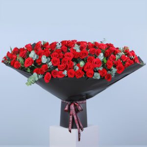 The image shows a large bouquet of red roses arranged in a black vase. The bouquet is wrapped in black paper with a red ribbon. The roses are interspersed with greenery, possibly eucalyptus. The arrangement appears to be quite grand and luxurious.